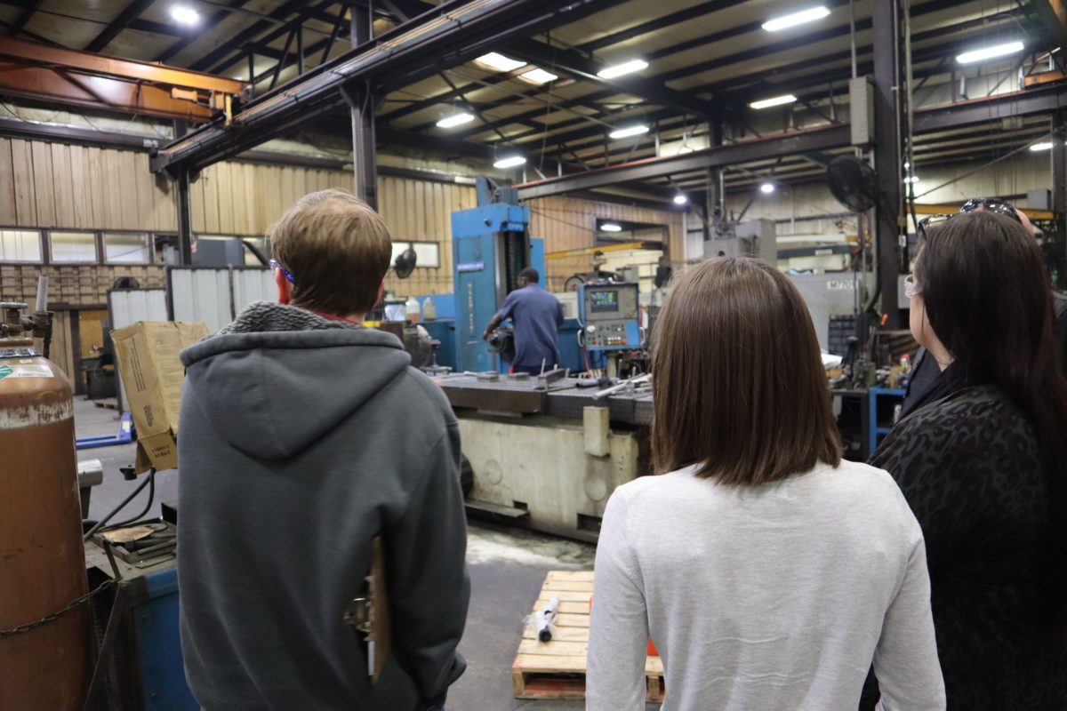 3 people with their backs to the camera, look at machines in a warehouse setting.