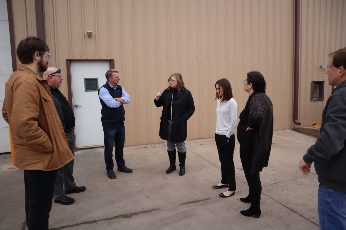 Group of 7 people standing outside of a metal building. Everyone is looking at the man in the middle who is speaking.